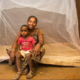 A woman and a young child sit in front of a mattress that has a mosquito net around it.