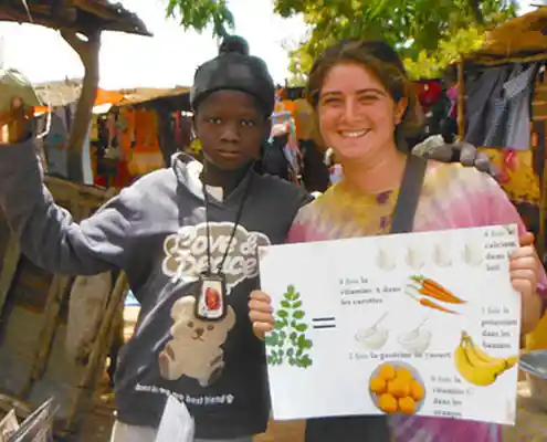 A Peace Corps worker and a young boy pose for the camera at an outdoor marketplace. The worker is holding up a poster for healthy foods.