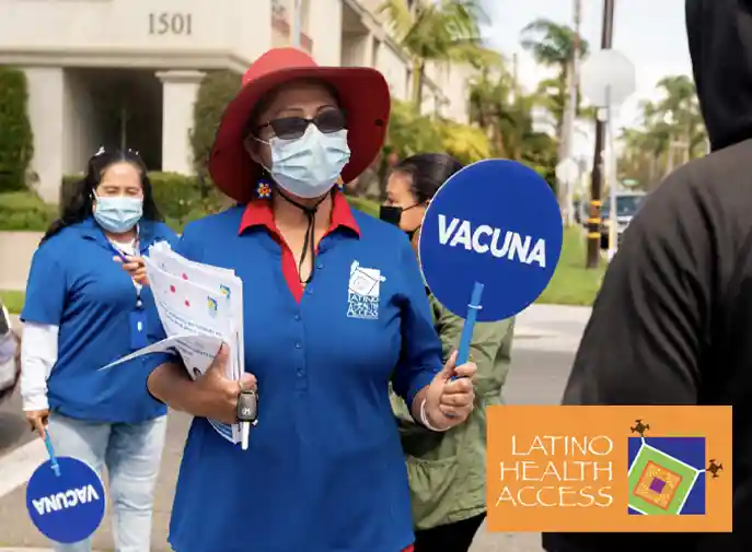 Two women wear masks and hold Vacuna signs as they try to encourage people to get vaccinated. Latino Health Access logo is at bottom right.