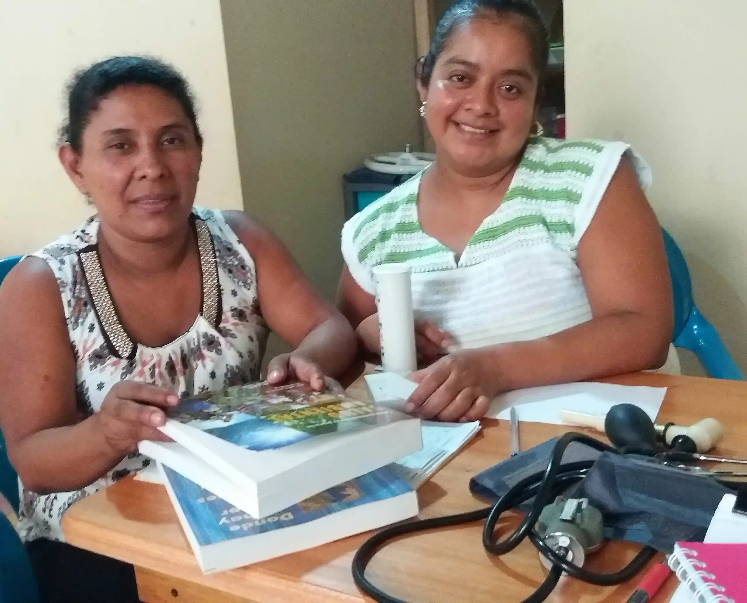 Two ASECSA workers sit at a desk and smile for the camera. Copies of Guía comunitaria para la salud ambiental and Donde no hay doctor sit on the desk in front of them. Stethoscopes are also on desk.