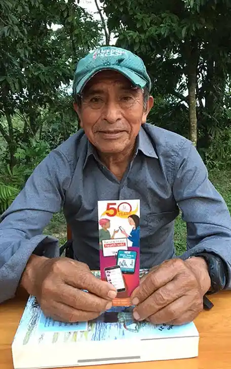 A older man smiles at camera while holding a Hesperian bookmark.