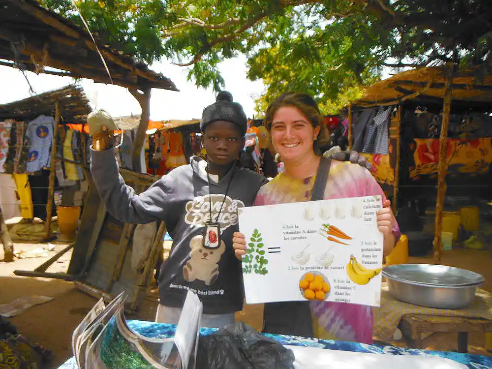 A peace corps volunteer and a young man holding up an apple in an African market place display a nutrition poster to the camera