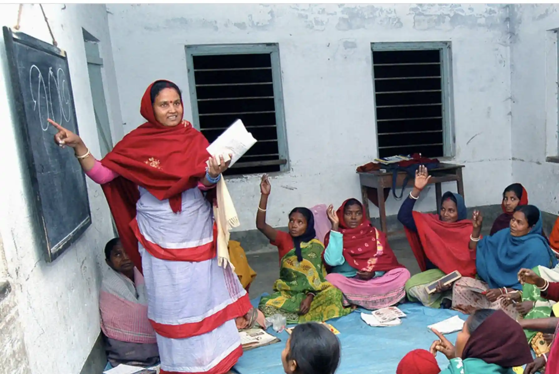 A health worker is giving a lesson in front of a chalkboard to a group of women. The women are sitting on the floor and some have their hands up.