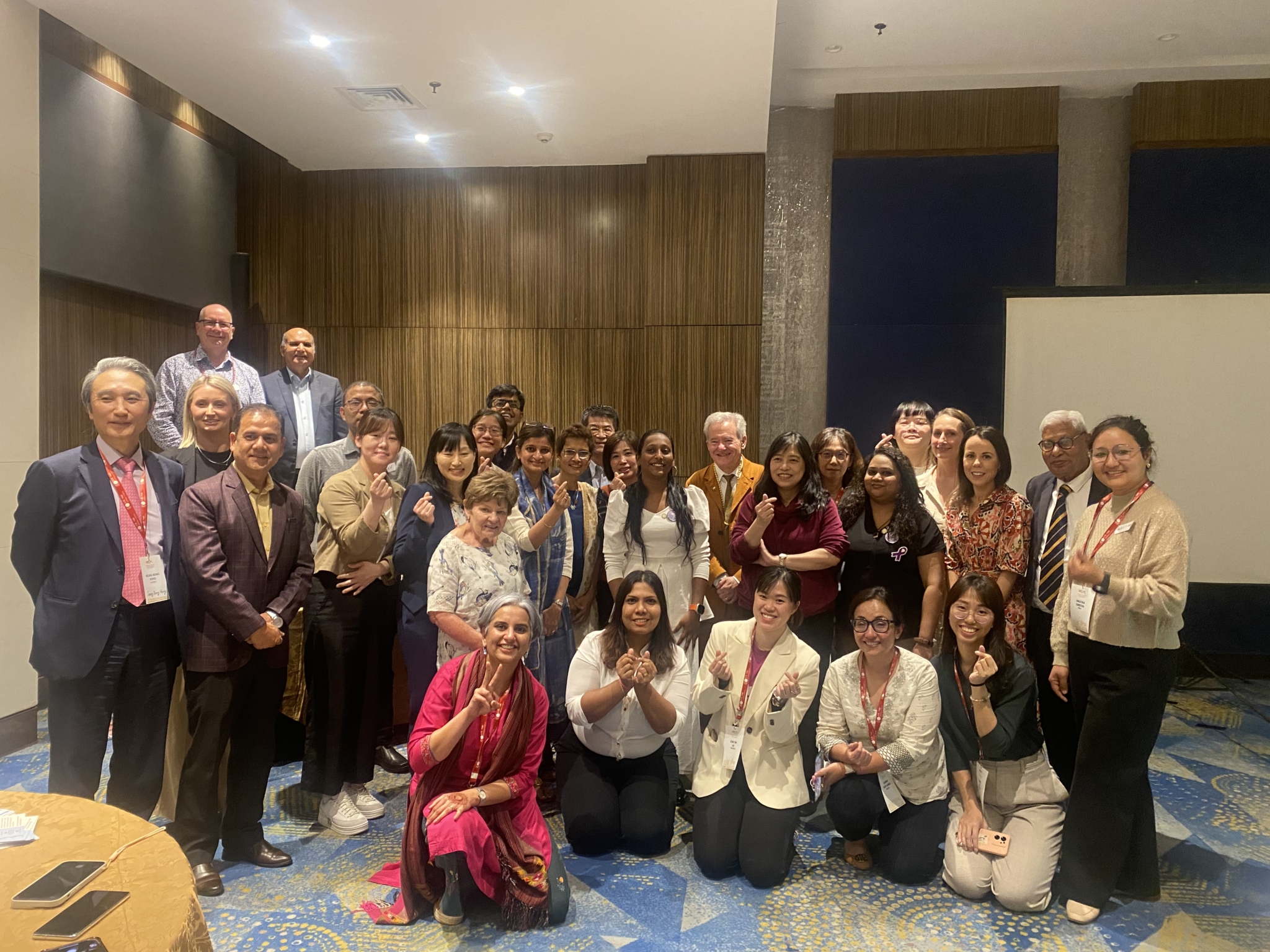 A group of men and women pose for a photo in a conference room. Several hold up their hands making peace signs and hearts